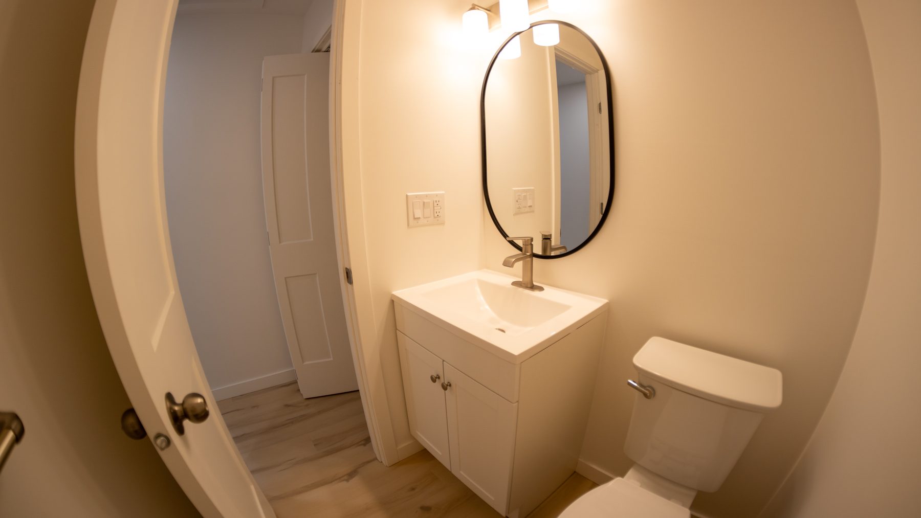 Finished powder room with white vanity, oval black-framed mirror and light wood-look flooring